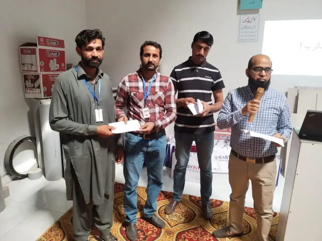An envelope handed to a man with a lanyard at a TurkPlast and Easyfit event, the host at the microphone